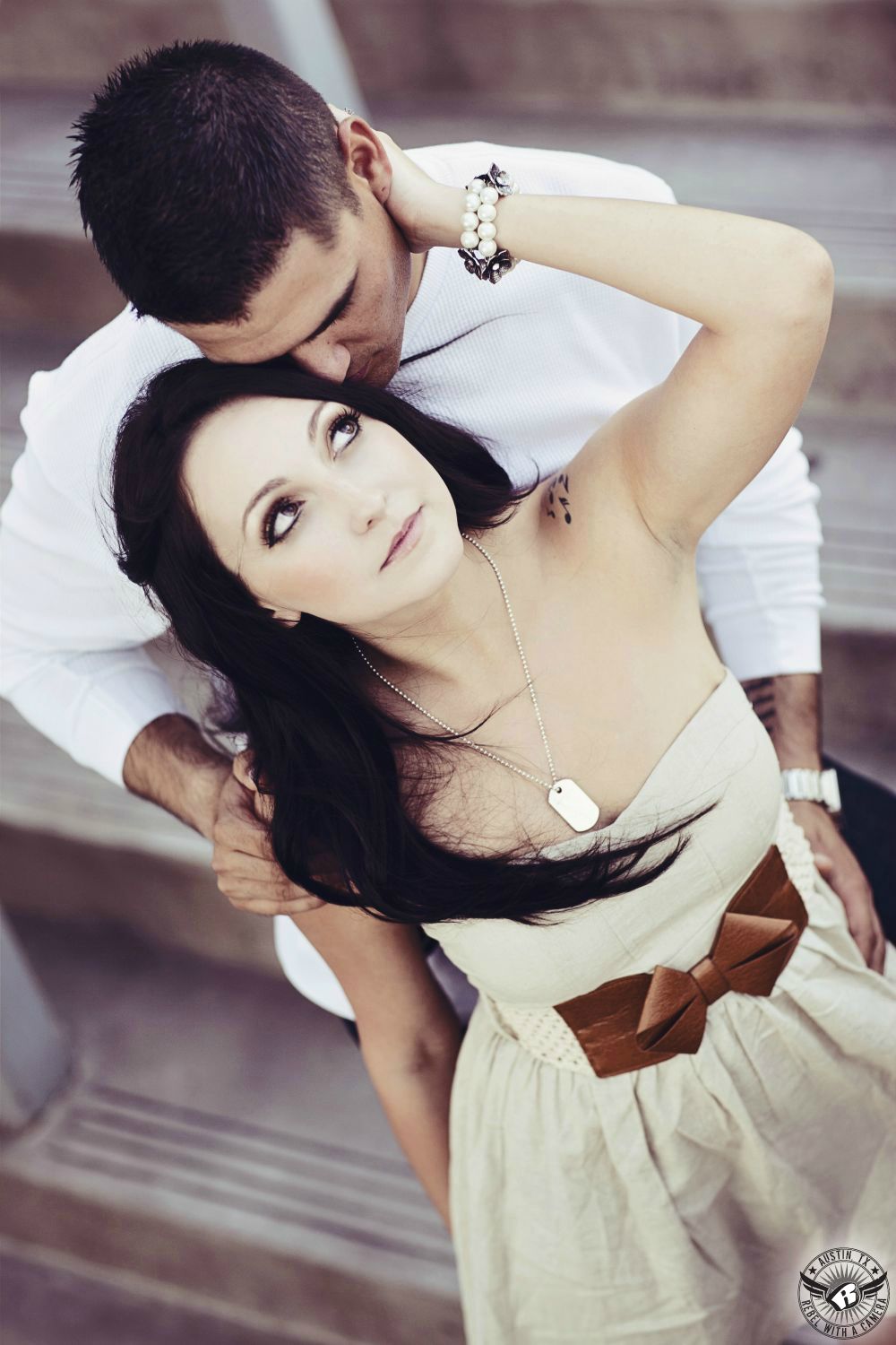 Dark haired Latino girl with light green strapless short dress with a golden bow and a dog tag necklace and pearls wristlet  looks up and a brunette guy wearing a white shirt with long sleeves as he nuzzles her hair on the front steps of The Long Center For Performing Arts in this enjoyable engagement portrait in downtown Austin.
